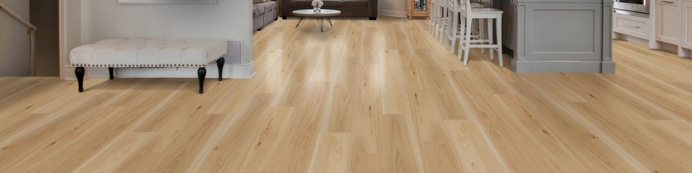 A light LVP wood floor extends through a home, with a white bench in the foreground and glimpses of a living area and kitchen island in the background.